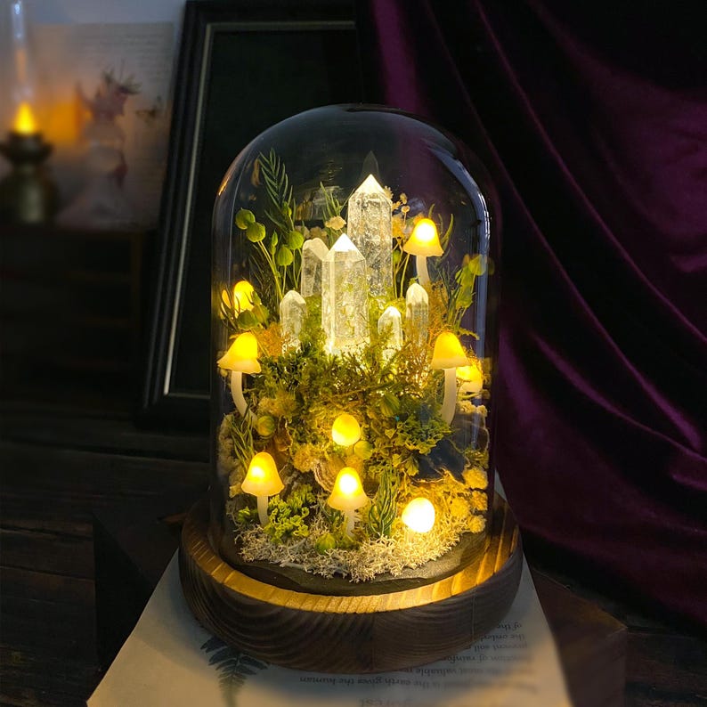 May include: A glass dome with a wooden base containing a miniature forest scene with white quartz crystals, green moss, and small yellow mushrooms. The dome is illuminated from within, creating a warm glow.