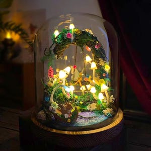 May include: A whimsical fairy garden scene encased in a glass dome. Miniature glowing mushrooms, flowers, and a fairy figurine are arranged within a mossy landscape. The dome sits on a wooden base, creating a magical display.