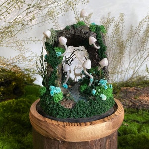May include: A miniature woodland scene with a small deer figurine, white mushrooms, and a stream in a wooden planter. The scene is surrounded by moss and other greenery.