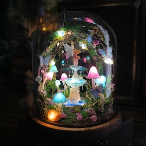 Peut inclure: Une sc&egrave;ne de jardin f&eacute;erique miniature enferm&eacute;e dans un d&ocirc;me en verre. Le paysage comprend une fontaine, des champignons lumineux, des papillons et une figurine de f&eacute;e. La sc&egrave;ne est &eacute;clair&eacute;e par de petites lumi&egrave;res LED, cr&eacute;ant une ambiance magique.