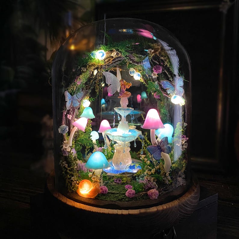 Peut inclure: Une sc&egrave;ne de jardin f&eacute;erique miniature enferm&eacute;e dans un d&ocirc;me en verre. Le paysage comprend une fontaine, des champignons lumineux, des papillons et une figurine de f&eacute;e. La sc&egrave;ne est &eacute;clair&eacute;e par de petites lumi&egrave;res LED, cr&eacute;ant une ambiance magique.