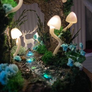 May include: A miniature forest scene with a white deer figurine, a stream with blue glitter, and glowing white mushrooms. The scene is set in a mossy, woodland environment.