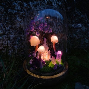 May include: A glass dome with a wooden base contains a miniature fairy garden with purple flowers, a small house, and glowing mushroom-shaped lights.