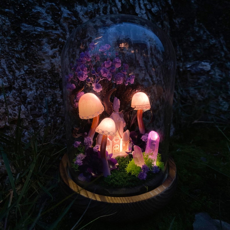 May include: A glass dome with a wooden base contains a miniature fairy garden with purple flowers, a small house, and glowing mushroom-shaped lights.