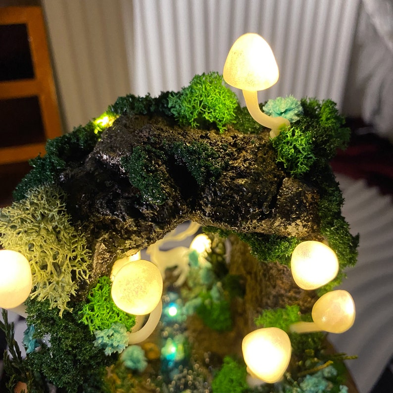 May include: A miniature forest scene with a mossy tree trunk and glowing white mushrooms. The scene is lit with small white lights, creating a magical and whimsical atmosphere.