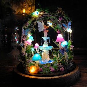 Peut inclure: Une sc&egrave;ne de jardin f&eacute;erique miniature dans un d&ocirc;me en verre. Elle comprend une petite fontaine, des champignons lumineux, des papillons et une figurine de f&eacute;e. La sc&egrave;ne est &eacute;clair&eacute;e par des lumi&egrave;res LED color&eacute;es, cr&eacute;ant une ambiance magique.