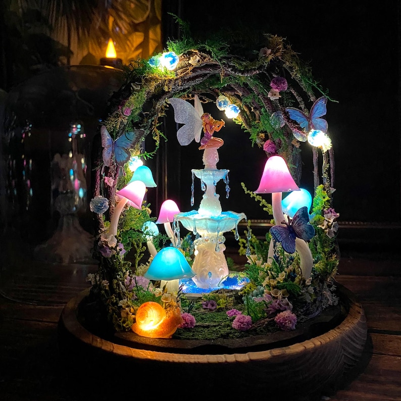 Peut inclure: Une sc&egrave;ne de jardin f&eacute;erique miniature dans un d&ocirc;me en verre. Elle comprend une petite fontaine, des champignons lumineux, des papillons et une figurine de f&eacute;e. La sc&egrave;ne est &eacute;clair&eacute;e par des lumi&egrave;res LED color&eacute;es, cr&eacute;ant une ambiance magique.