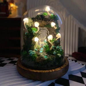 May include: A glass dome terrarium with a miniature forest scene. The scene includes a small deer figurine, a stream of iridescent blue stones, and a variety of green moss and plants. The dome is lit by small white LED lights shaped like mushrooms.