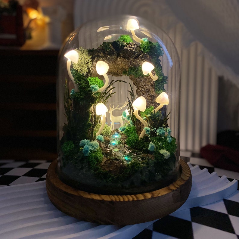 May include: A glass dome terrarium with a miniature forest scene. The scene includes a small deer figurine, a stream of iridescent blue stones, and a variety of green moss and plants. The dome is lit by small white LED lights shaped like mushrooms.