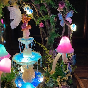 Peut inclure: Une sc&egrave;ne de jardin f&eacute;erique fantaisiste avec une fontaine lumineuse, une figurine de f&eacute;e, des champignons et des papillons. La fontaine a de l'eau en cascade et est entour&eacute;e de verdure et de fleurs. La sc&egrave;ne est &eacute;clair&eacute;e par des lumi&egrave;res douces et color&eacute;es.