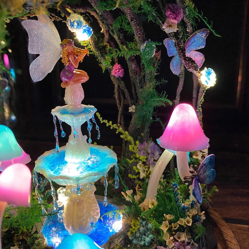 Peut inclure: Une sc&egrave;ne de jardin f&eacute;erique fantaisiste avec une fontaine lumineuse, une figurine de f&eacute;e, des champignons et des papillons. La fontaine a de l'eau en cascade et est entour&eacute;e de verdure et de fleurs. La sc&egrave;ne est &eacute;clair&eacute;e par des lumi&egrave;res douces et color&eacute;es.