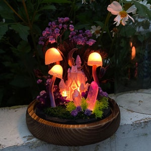 May include: A miniature fairy tale castle with glowing lights sits in a wooden planter surrounded by purple flowers, moss, and crystals. Three mushroom-shaped lamps illuminate the scene.