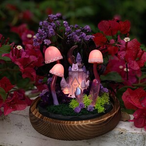 May include: A miniature fairy tale castle with pink and purple accents sits in a wooden planter with pink mushrooms, purple crystals, and green moss. The castle has a crescent moon on top and a heart-shaped window.