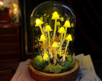 May include: A decorative glass dome featuring illuminated yellow mushroom lights. The mushrooms are arranged with moss and dried flowers on a wooden base. The dome creates a warm, inviting glow, perfect for home decor.