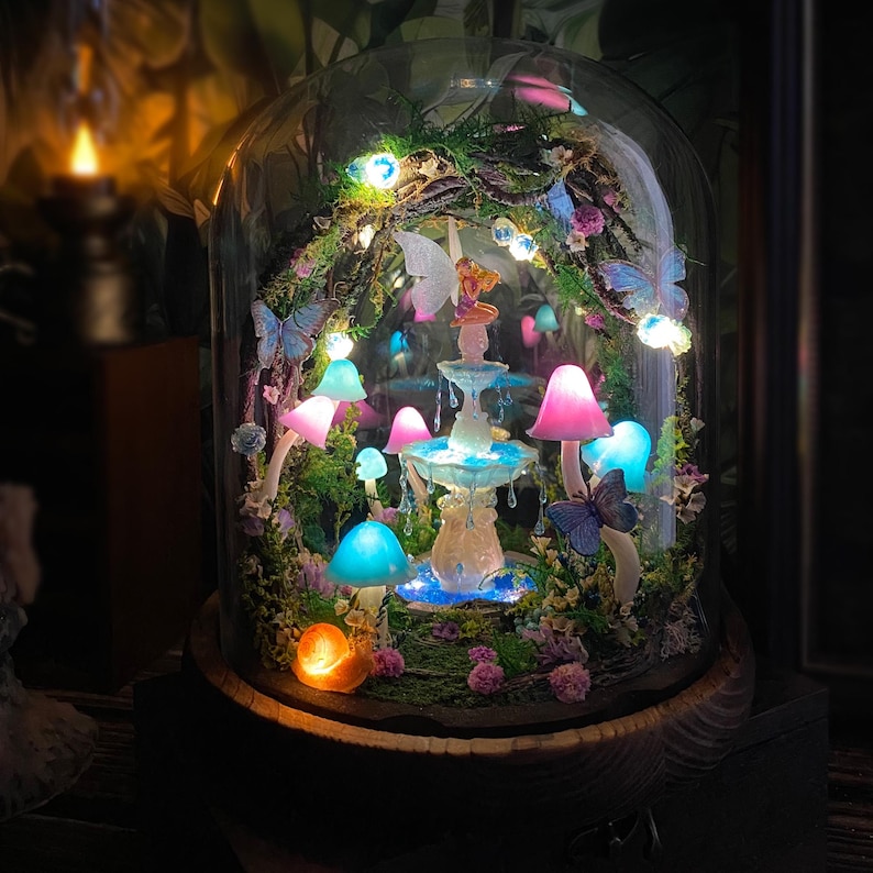 Peut inclure: Une sc&egrave;ne f&eacute;erique fantaisiste sous un d&ocirc;me de verre. Le paysage miniature comprend des champignons lumineux aux chapeaux roses et bleus, une fontaine, des papillons et une figurine de f&eacute;e. La base est en bois sombre, et la sc&egrave;ne est &eacute;clair&eacute;e par des guirlandes lumineuses.