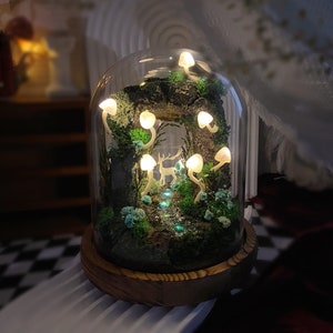 May include: A glass dome with a wooden base contains a miniature forest scene with glowing white mushrooms and a white deer figurine. The forest is made of moss and branches, and there is a small stream of water flowing through it.
