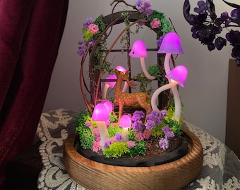 Può includere: Una base in legno con una scena di foresta in miniatura che presenta un cervo, muschio e funghi viola con luci brillanti. La scena è racchiusa in una cornice in legno con una finestra.