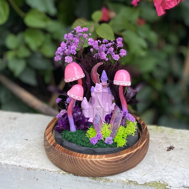 May include: A miniature fairy garden scene with a pink castle, pink mushrooms, purple flowers, and iridescent crystals in a wooden planter.