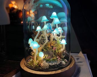 May include: A glass dome with a wooden base containing a miniature fairy garden with glowing blue mushrooms and a small fairy figurine.