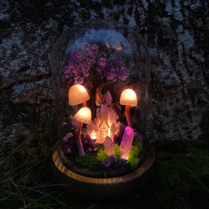 May include: A glass dome with a miniature fairy house, purple flowers, and glowing mushrooms. The house has a crescent moon above it and is surrounded by crystals.