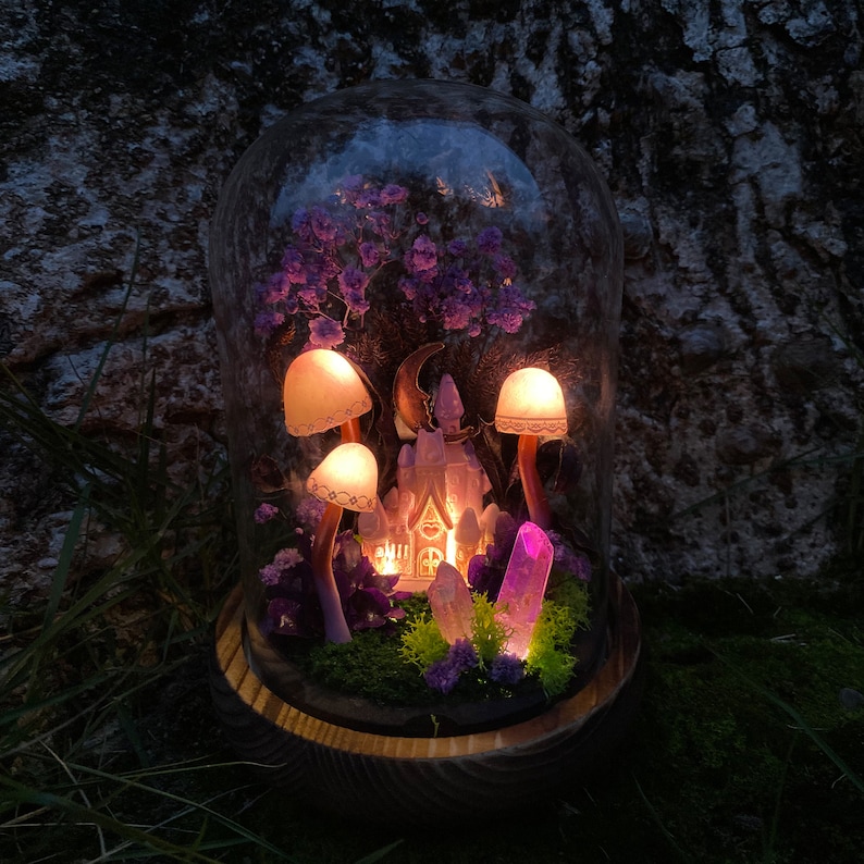 May include: A glass dome with a miniature fairy house, purple flowers, and glowing mushrooms. The house has a crescent moon above it and is surrounded by crystals.