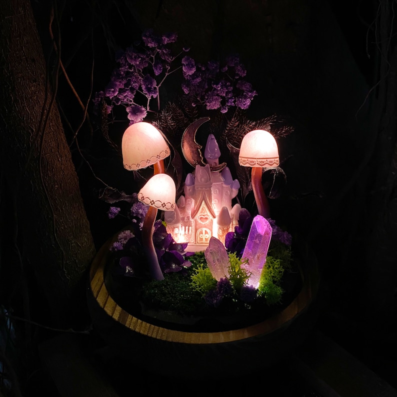 May include: A miniature fairy house with a heart-shaped window, surrounded by purple flowers, crystals, and glowing mushroom lamps in a wooden planter.