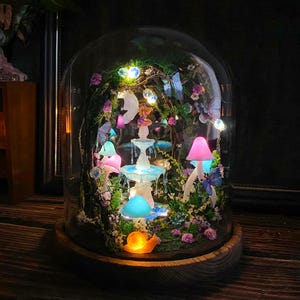Peut inclure: Une sc&egrave;ne de jardin f&eacute;erique miniature enferm&eacute;e dans un d&ocirc;me en verre. Le paysage miniature comprend une fontaine lumineuse, des champignons color&eacute;s, des papillons et un escargot. La base est en bois et la sc&egrave;ne est &eacute;clair&eacute;e par des lumi&egrave;res douces et chaudes.