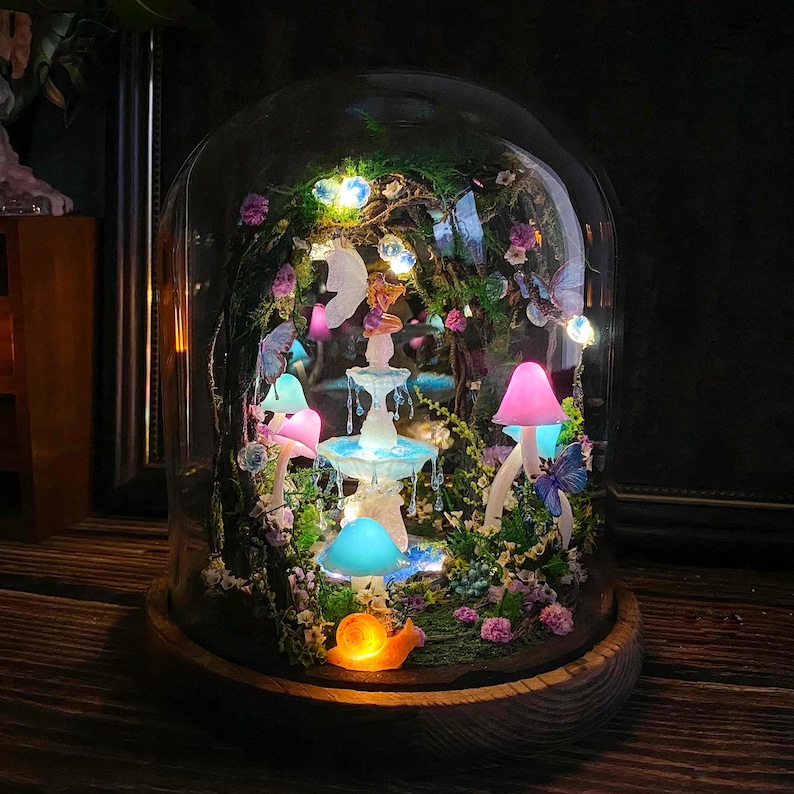 Peut inclure: Une sc&egrave;ne de jardin f&eacute;erique miniature enferm&eacute;e dans un d&ocirc;me en verre. Le paysage miniature comprend une fontaine lumineuse, des champignons color&eacute;s, des papillons et un escargot. La base est en bois et la sc&egrave;ne est &eacute;clair&eacute;e par des lumi&egrave;res douces et chaudes.