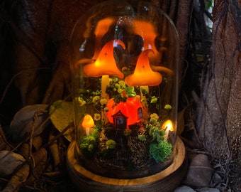 Puede incluir: Una campana de cristal con una escena de bosque en miniatura en su interior. La escena incluye una pequeña casa naranja, musgo verde y velas en forma de hongo naranja. La campana está sentada sobre una base de madera.