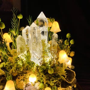 May include: A crystal cluster featuring clear quartz points, surrounded by small, glowing, yellow mushroom-shaped lights. The crystal cluster is nestled in a bed of green moss and other foliage.