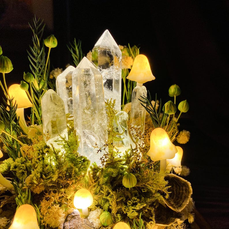 May include: A crystal cluster featuring clear quartz points, surrounded by small, glowing, yellow mushroom-shaped lights. The crystal cluster is nestled in a bed of green moss and other foliage.