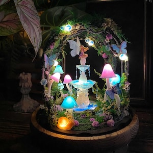 Peut inclure: Une sc&egrave;ne de jardin f&eacute;erique miniature avec une fontaine lumineuse, des champignons color&eacute;s et des papillons. La sc&egrave;ne est &eacute;clair&eacute;e par de petites lumi&egrave;res, cr&eacute;ant un affichage fantaisiste. La base est un bol en bois.