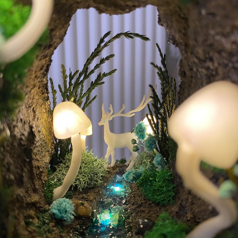 May include: A miniature forest scene with a white deer figurine, a small stream, and glowing white mushrooms. The scene is set inside a rock cave with green foliage.