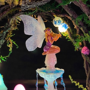 Peut inclure: Une figurine de f&eacute;e miniature jouant du violon, perch&eacute;e au sommet d'une fontaine lumineuse. La f&eacute;e a des cheveux blonds, une robe violette et des ailes iris&eacute;es. La fontaine a des accents bleus et blancs, avec des gouttelettes d'eau en cascade. La sc&egrave;ne se d&eacute;roule dans un environnement fantaisiste et &eacute;clair&eacute;.
