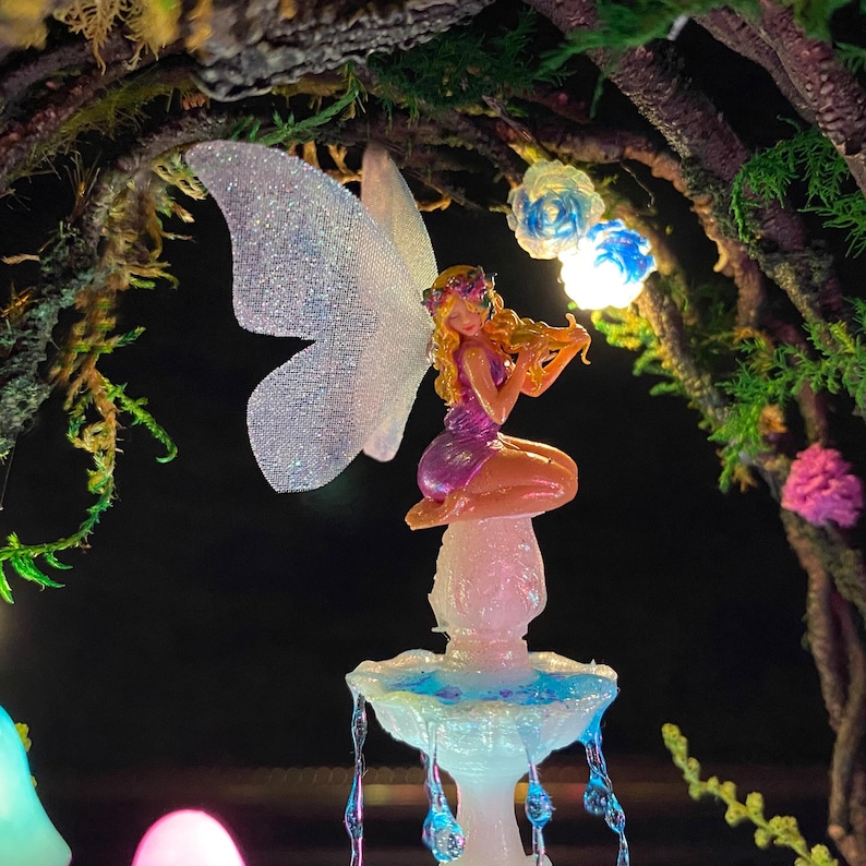 Peut inclure: Une figurine de f&eacute;e miniature jouant du violon, perch&eacute;e au sommet d'une fontaine lumineuse. La f&eacute;e a des cheveux blonds, une robe violette et des ailes iris&eacute;es. La fontaine a des accents bleus et blancs, avec des gouttelettes d'eau en cascade. La sc&egrave;ne se d&eacute;roule dans un environnement fantaisiste et &eacute;clair&eacute;.