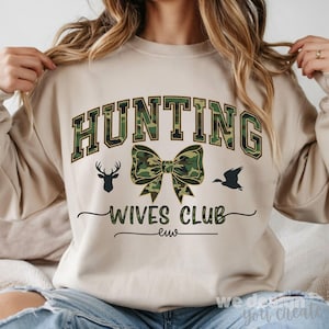 以下が含まれることがあります： ベージュのスウェットシャツ。テキスト「HUNTING WIVES CLUB」は黒と迷彩グリーン。デザインには、リボン、鹿のシルエット、飛んでいるアヒルのシルエットが含まれています。