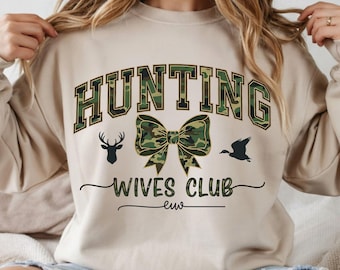 Op de afbeelding: Beige sweatshirt met de tekst "HUNTING WIVES CLUB" in zwart en camouflagegroen. Het ontwerp bevat een strik, een herten silhouet en een vliegend eend silhouet.