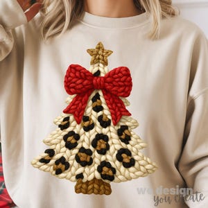 May include: Cream-colored sweatshirt featuring a knitted Christmas tree design. The tree has a red bow, gold star, and leopard print spots. The text "we design you create" is at the bottom right.