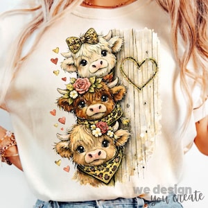 Può includere: T-shirt color crema con un design a strati di tre mucche dei cartoni animati. Le mucche hanno accenti floreali e animalier. È visibile uno sfondo di assi di legno con un contorno a forma di cuore. Il testo "we design you create" è in basso.