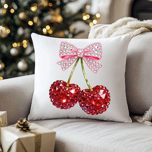 May include: White decorative pillow featuring a pair of red jewel-encrusted cherries with green stems and a pink jeweled bow. The pillow is on a gray couch with a Christmas tree in the background.