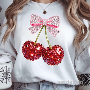 May include: White sweatshirt featuring a sparkling pink bow and two red cherries with a jewel-like design. The cherries have green stems. The text "we design you" is at the bottom right.