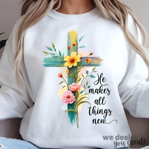 May include: White sweatshirt with a teal cross and floral design. The text "He makes all things new" is printed on the sweatshirt.
