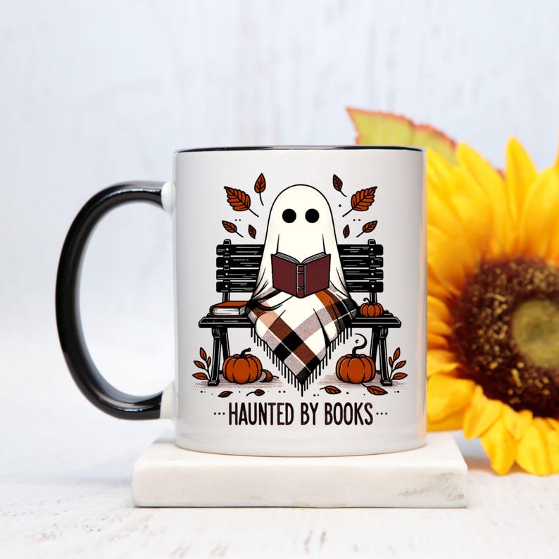 以下が含まれることがあります： 黒いハンドルと縁が付いた白いセラミックマグ。マグには、カボチャと秋の葉に囲まれた、本を読む幽霊が描かれています。マグのテキストは「...HAUNTED BY BOOKS...」と書かれています。