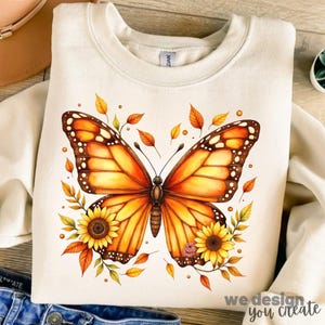 May include: Cream-colored sweatshirt featuring a large, detailed butterfly design in shades of orange, yellow, and brown. The butterfly is surrounded by autumn leaves and sunflowers. The text "we design you create" is at the bottom right.
