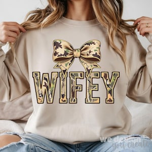 以下が含まれることがあります： ベージュのスウェットシャツ。大きなリボンと「WIFEY」の文字が迷彩柄でデザインされています。リボンは茶色、緑、ベージュ。文字は濃い茶色で縁取られています。
