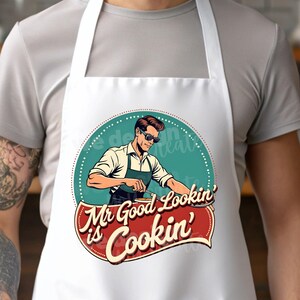 Peut inclure: Tablier blanc avec un design r&eacute;tro repr&eacute;sentant un homme portant des lunettes de soleil et un tablier, cuisinant sur un barbecue. Le texte dit "Mr Good Lookin' is Cookin'".