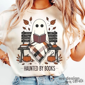 以下が含まれることがあります： 公園のベンチで本を読む漫画の幽霊が描かれたクリーム色のTシャツ。幽霊はチェック柄の毛布に包まれ、カボチャと秋の葉に囲まれています。「...HAUNTED BY BOOKS...」の文字が下に印刷されています。