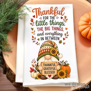 May include: A white kitchen towel featuring a gnome wearing a patterned hat with a sunflower. The text reads: Thankful for the little things, the big things, and everything in between. The gnome holds a sign saying: Thankful, Grateful, Blessed.