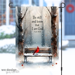 Könnte beinhalten: Eine Gartenflagge mit einer Winterlandschaft, einem roten Kardinal auf einer Bank. Die Flagge trägt den Text "Be still and know that I am God, Psalm 46:10". Das Design ist im Aquarellstil gehalten und enthält den Text "we design you create."