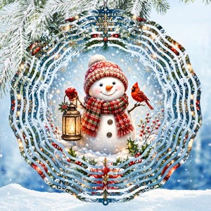 May include: A circular metal wind spinner featuring a snowman wearing a red patterned hat and scarf, holding a lit lantern. A red cardinal sits on a branch beside the snowman, surrounded by holly berries and snowflakes against a blue winter background.